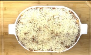 Baked cheesy casserole in a white dish on a wooden cutting board, topped with shredded cheese.